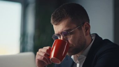 Modern bir ofiste dizüstü bilgisayarla çalışırken gözlüklü bir adam kahvesini yudumluyor. İş ortamında konsantrasyon, verimlilik ve profesyonel odaklanma kavramı.