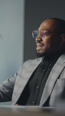 Dikey: Modern bir ofiste Afro-Amerikalı profesyonel kahve yudumluyor, bir masada dizüstü bilgisayarla oturuyor ve arka planda bitkiler var. İş yeri rutini, odaklanma ve rahatlama kavramı.