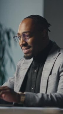 Dikey Ekran: Modern bir ofiste dizüstü bilgisayarda çalışan, ekose takım elbiseli ve gözlüklü kendinden emin Afro-Amerikan bir adam. Üretkenlik kavramı, iş yeniliği, işyeri odağı.