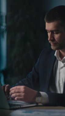 Dikey Ekran: Düşünceli iş adamı gece geç saatlere kadar bir ofiste çalışır, dizüstü bilgisayar kullanırken yorgunluk ve düşünce anlarını gösterir. İş yeri stresi ve mesleki zorluklar kavramı.