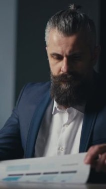 Dikey Ekran: Profesyonel yönetici loş bir ofiste masada oturuyor, akıllı telefonunu kontrol ediyor ve belgeleri inceliyor. Odaklanma, teknoloji ve modern iş anlayışı.