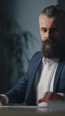 Dikey Ekran: İşadamı geç saatlere kadar masasında çalışıyor, modern ofis ortamında bir dizüstü bilgisayardaki belgeleri inceliyor. Kurumsal bir ortamda odaklanma, kararlılık ve profesyonellik kavramı.