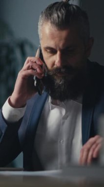 Dikey Ekran: Girişimci ofiste geç saatlere kadar çalışıyor, belgelere bakarken telefonla konuşuyor ve dizüstü bilgisayar kullanıyor. Profesyonel iletişim, çoklu görev ve kurumsal çalışma kavramı.