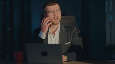 Takım elbiseli bir iş adamı, geceleri telefonla meşgul, loş bir ofiste dizüstü bilgisayarla çalışıyor. Geç saatlere kadar çalışma, iş görüşmeleri ve şirket stratejisi kavramı.