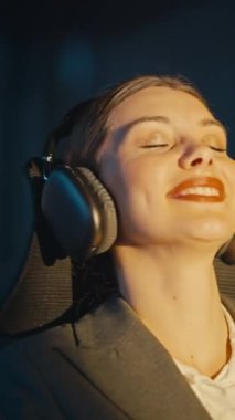 Dikey Ekran: Takım elbiseli şık bir kadın büyük kulaklıklı müzikten hoşlanıyor, gülümseyerek arkasına yaslanıyor. Bir iş ortamında rahatlama, müzik takdir ve stres rahatlama kavramı.