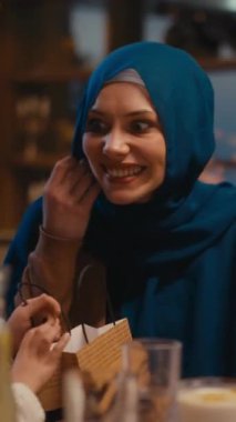 Dikey Ekran: Mavi tesettürlü Müslüman kadın, çocuk iftar sırasında veya bayram yemeğinde hediye alırken neşeli bir anı paylaşıyor. Aile bağı, cömertlik, kültürel gelenek kavramı