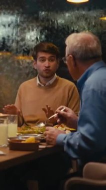 Dikey ekran: yaşlı bir adam ve genç bir adam bir restoranda yemek yerken sohbet ediyorlar, sıcak bir ortamda yemek yiyorlar. Nesiller arası diyalog kavramı, iftar ve bağ.