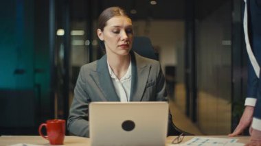 Gri takım elbiseli profesyonel bir kadın loş bir ofiste geç saatlere kadar çalışır, erkek bir meslektaşı ona yaklaşırken ve elini omzuna koyarken stres ve rahatsızlık gösterir. İş yeri tacizi kavramı.