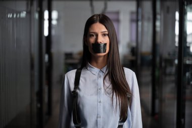 Ciddi bir kadın, ağzında siyah bantla ofiste duruyor. Sansürü, baskıyı ya da işyeri ayrımcılığını simgeliyor. Konuşma özgürlüğü kavramı, işyeri eşitsizliği veya baskı.