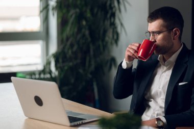 Gözlüklü profesyonel bir adam, takım elbise giyiyor, geç saatlere kadar masa başında kahve içiyor. Odaklanma, üretkenlik ve modern ofis yaşam tarzı kavramı.