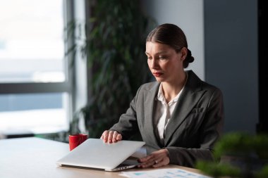Modern bir ofisteki iş kadını laptopunu açıyor, odaklanma ve hafif kaygı karışımıyla çalışmaya hazırlanıyor. Mesleki zorluklar kavramı, kurumsal nişanlar, son teslim tarihinden endişe duymak..