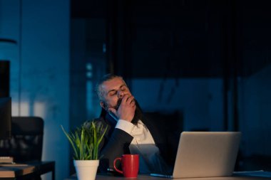 Gece geç saatlere kadar çalışan yorgun girişimci, dizüstü bilgisayarla aydınlatılmış loş bir ofiste esniyor. Aşırı çalışma, gece yarısı üretkenliği, yorgunluk ve iş-yaşam dengesi kavramı.