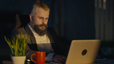 Gece geç saatlere kadar loş bir ofiste çalışan bir iş adamı laptopuna odaklanmış. Bağlılık, fazla mesai, kurumsal sorumluluk, iş stratejisi ve üretkenlik kavramı.