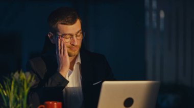 Karanlık bir ofiste gece geç saatlere kadar çalışan yorgun iş adamı dizüstü bilgisayar ekranına bakarken yüzünü yorgunlukla ovuyor. Aşırı çalışma, stres ve kurumsal tükenme kavramı.