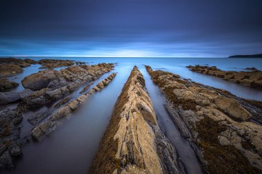 Huzurlu bir deniz manzarası çarpıcı bir alacakaranlık gökyüzünün altında engebeli kayalık sahili yakalıyor.