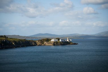 Bu sahne, berrak mavi gökyüzünün altında yuvarlanan tepelerin yanında büyüleyici bir deniz feneri gösteren nefes kesici güzellikte bir kıyı manzarası yakalar.
