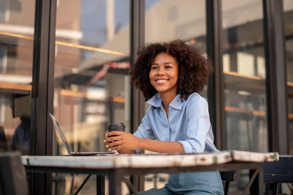 Elinde kahve fincanı olan güzel bir Afrikalı kadın laptopuyla kafenin dışında oturuyor.