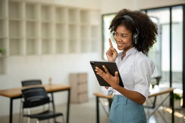 African american college student with headphones in hand with tablet standing smiling. Look at a specific point with your finger pointing up. in cafe or workspace