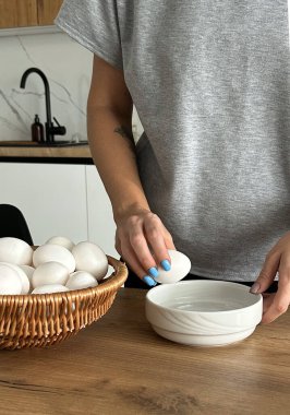 Yakın çekimde, kadın bir aşçı ev mutfağındaki ahşap bir masada yemek hazırlamak için bir fincana yumurta kırıyor. Yumurta sarısı yemek sağlıklı bir kahvaltıdır..