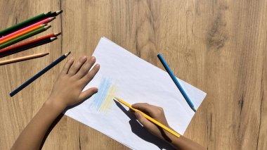 Çocuk çiziyor. Bir çocuk Ukrayna bayrağını çekiyor. Ahşap bir masa üzerinde renkli kalemler ve beyaz kağıt, üst manzara.