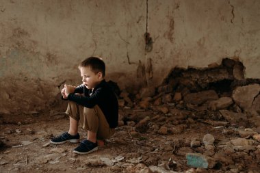 Üzgün, küçük bir Ukraynalı çocuk, bir anaokulu öğrencisi, yıkılmış evinin yıkıntıları üzerinde oturuyor ve düşünüyor.