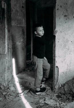 Siyah tişörtlü bir çocuk kapının yanında duruyor ve harap olmuş evinden pencereden dışarı bakıyor. Ukrayna 'da savaş. Savaşın sonuçları. Siyah beyaz fotoğraf.
