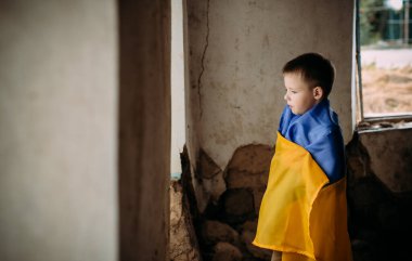 Bir çocuk harap bir evde duruyor ve pencereden dışarı bakıyor. Ukrayna bayrağına sarılmış bir çocuk harabelerde duruyor. Ukrayna 'da savaş. Savaşın sonuçları..