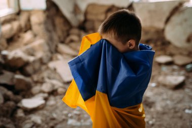 Bir çocuk harap olmuş bir evde durur ve yüzünü kapatarak ağlar. Harabelerin arasında Ukrayna bayrağına sarılmış bir çocuk duruyor. Ukrayna 'da savaş. Savaşın sonuçları..