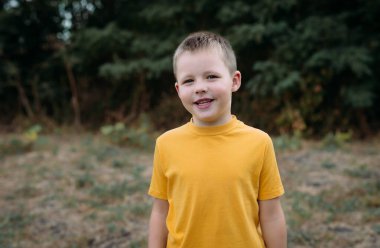 Küçük mutlu bir çocuk sokakta sarı bir tişörtle dikiliyor. Bir çocuk parkta yürürken mutlu bir şekilde gülümsüyor. Neşeli çocukluk kavramı.