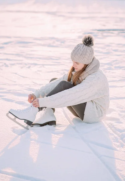 Young woman ice skating on a frozen lake in a snowy winter park during sunset. A girl sits on the ice tying her skates before skating at the skating rink. Side view. Active recreation in winter.