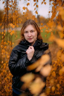 Renkli sonbahar yapraklarının yanında duran koyu renk ceketli güzel bir kızın portresi. Genç bir kadın, sonbahar mevsiminde sarı ağaçların arka planında parkta dinlenir..