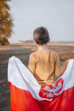 Arkasında Polonya bayrağını tutan yakışıklı küçük bir çocuk. Doğada ulusal bayrak taşıyan bir çocuk yolda duruyor. Ulusal bayramlar ve vatanseverlik olayları kavramı. Bağımsızlık Günü