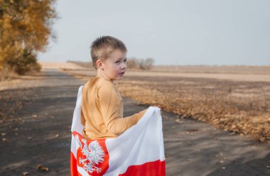 Arkasında Polonya bayrağını tutan yakışıklı küçük bir çocuk. Doğada ulusal bayrak taşıyan bir çocuk yolda duruyor. Ulusal bayramlar ve vatanseverlik olayları kavramı. Bağımsızlık Günü