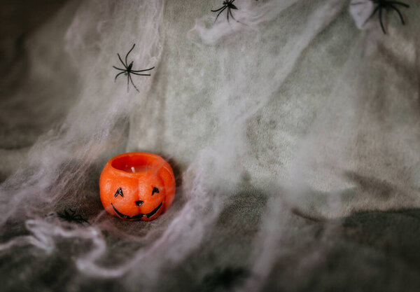 Halloween at home, room decoration ideas. A wax candle in the shape of a smiling pumpkin in webs and spiders. Symbol of All Saints' Day, October holidays. Gray background.