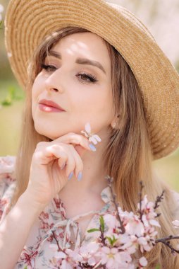 Renkli desenli beyaz elbiseli bir kadın gözleri kapalı poz veriyor. Şapkalı güzel bir genç kadın bahçede çiçek açan bir bahar ağacının yanında duruyor ve elinde bir çiçek tutuyor..