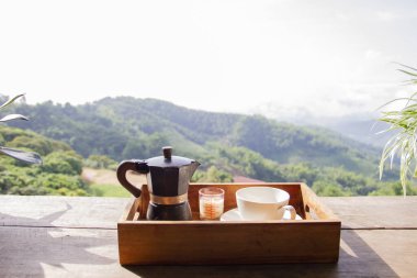 Kahve makinesi seti, ahşap kutunun üzerinde moka demliği ve arka plan olarak dağ ve köy.