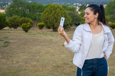 Güzel ve doğal Latin kadın parkta cep telefonuyla selfie çekiyor..