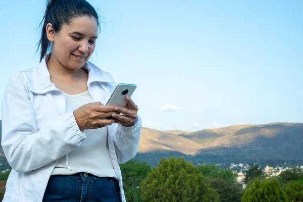 Güzel ve doğal Latin bir kadın parkta cep telefonunu kullanıyor. Parkta rahatlayıp cep telefonundan mesaj okuyan genç bir kadının portresi..
