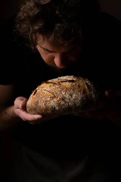 Attractive young Caucasian chef posing with white sourdough bread. The sourdough bread is the central protagonist of the scene, standing out with beautiful golden tones against the dark background.