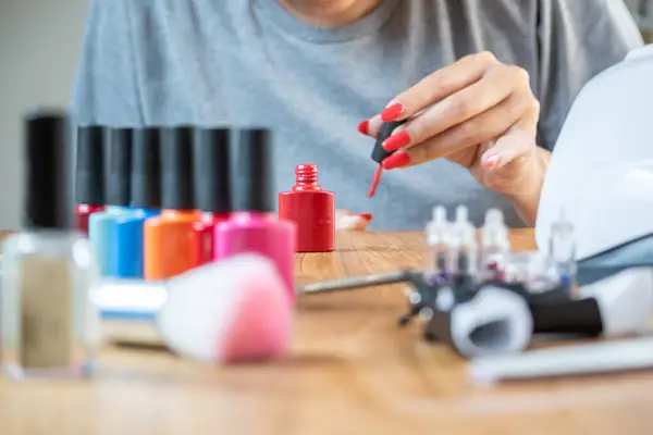 Woman paints her nails at the table at home. She has many nail polish colors to choose from.