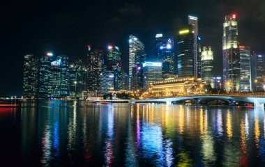 Singapur 'un ışıl ışıl silueti, güzel aydınlatılmış bir köprünün altında parıldayan rengarenk yansımalarıyla geceleri parlıyor..