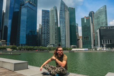 Singapur 'da bir turist, yüksek gökdelenlere ve berrak mavi gökyüzüne bakarken kıyıların yanında oturup dinleniyor..