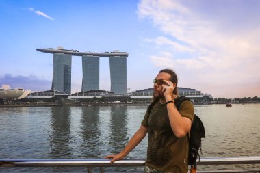 Suyun kenarında duran bir turist, arka plandaki ikonik Marina Bay Sands binasıyla telefon görüşmesi yapıyor..