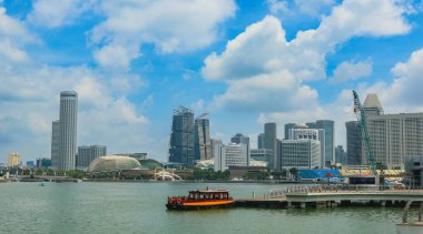 Singapur 'un gökdelenleri modern gökdelenler ve su üzerinde bir tekne, hepsi de kabarık bulutlu parlak mavi bir gökyüzüne karşı kurulu..