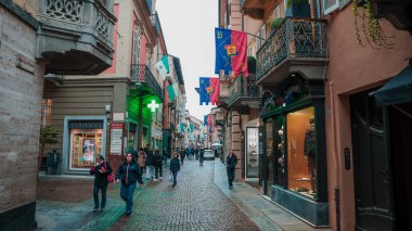 Alba, İtalya - 27 Ekim 2024: Alba İtalya 'da renkli bayraklarla süslenmiş canlı bir cadde. İnsanlar kaldırım taşları yolunda yürüyor, yerel dükkanların ve gri gökyüzünün altındaki canlı atmosferin tadını çıkarıyorlar..