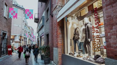 Alba, İtalya - 27 Ekim 2024: Ziyaretçiler İtalya 'nın Alba kentindeki büyüleyici bir ara sokakta dolaşırken, renkli afişler havada süzülürken, keşif ve alışverişe davetiye çıkaran canlı bir atmosfer yaratıyorlar..