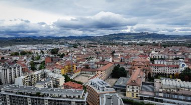 İtalya 'nın Alba şehrinin ufuk çizgisi, bulutlu bir gökyüzü altında tarihi mimari ve modern yapıların bir karışımını ortaya koyuyor. Uzakta kayan tepeler çarpıcı bir zemin sağlıyor..