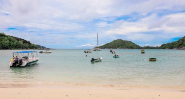 Seyşeller 'in sakin sularının tadını çıkaran ziyaretçiler, bulutlu bir gökyüzünün altında tekneler kıyıda huzur içinde yüzüyor..