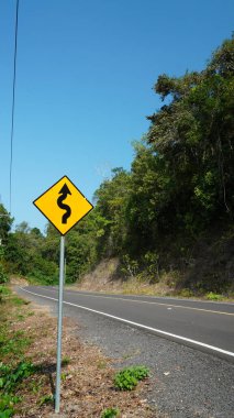 Manzara fotoğrafında, sürücülere çevrelerinin farkında olmaları ve hızlarını buna göre ayarlamaları gereğini vurgulayan dolambaçlı bir orman yolu için bir trafik tabelası yer alıyor..