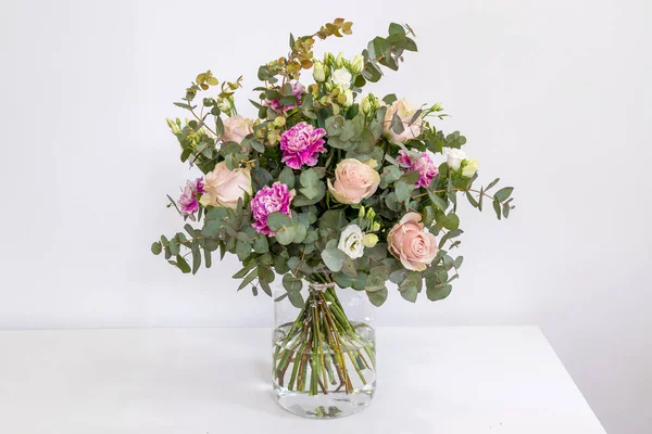 Bouquet of champagne roses, carnations and eucalyptus in a glass vase on a white background.